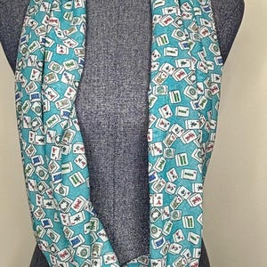 Op de afbeelding: Een turquoise oneindige sjaal met een witte achtergrond en een patroon van mahjong-tegels. De tegels zijn willekeurig over de sjaal verspreid.