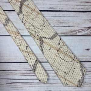 May include: A beige necktie with a brown grid pattern and brown line drawings resembling an EKG. The tie is made of a smooth fabric and is displayed on a light wood surface. The tie has a classic pointed end.