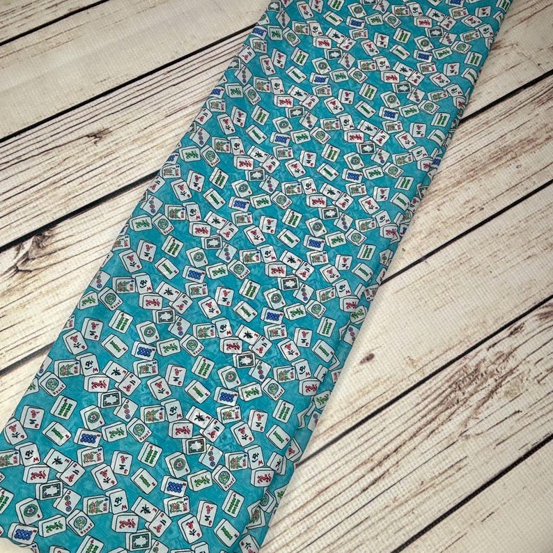 Op de afbeelding: Turquoise stof met een herhalend patroon van witte mahjong-tegels. De tegels hebben zwarte en rode Chinese karakters en symbolen.