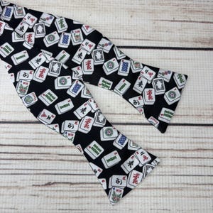 May include: A black bow tie featuring a repeating pattern of white Mahjong tiles with red, green, and blue accents. The bow tie is made of fabric and is designed to be tied around the neck. The bow tie is a fashion accessory.
