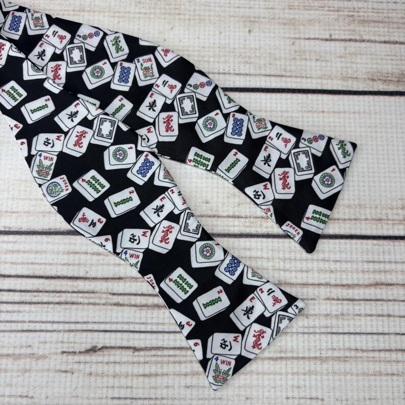 May include: A black bow tie featuring a repeating pattern of white Mahjong tiles with red, green, and blue accents. The bow tie is made of fabric and is designed to be tied around the neck. The bow tie is a fashion accessory.
