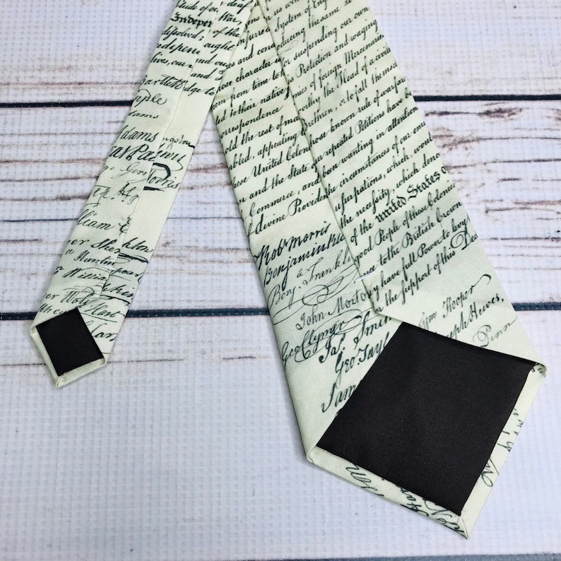 May include: Cream-colored necktie featuring a handwritten script design in black ink. The tie has a classic shape with a pointed end and a black backing. The script appears to be a historical document.