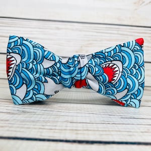 May include: A blue and white bow tie with a shark and wave pattern. The shark has red teeth and a white belly.