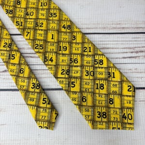 May include: A yellow necktie with a repeating pattern of a measuring tape design. The tie features black numbers and markings, creating a unique and eye-catching accessory. The tie is made of a smooth fabric.