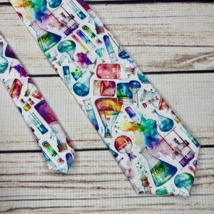 Könnte beinhalten: Eine weiße Krawatte mit einem Aquarelldesign aus farbenfrohen Laborgeräten, darunter Bechergläser, Kolben und Reagenzgläser. Die Krawatte hat ein sich wiederholendes Muster aus roten, blauen, grünen und gelben Elementen.