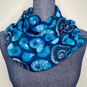 May include: A blue silk scarf with a pattern of white nautilus shells. The scarf is folded and displayed on a mannequin.