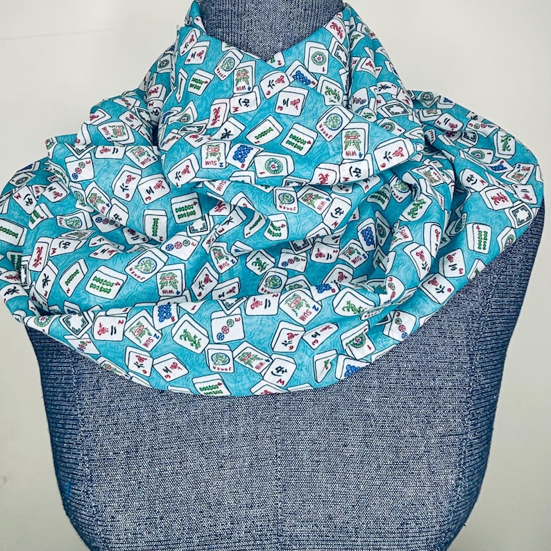 Op de afbeelding: Een turquoise blauwe sjaal met een witte achtergrond en een patroon van mahjong-tegels. De tegels zijn willekeurig over de sjaal verspreid en bevatten Chinese karakters en symbolen.
