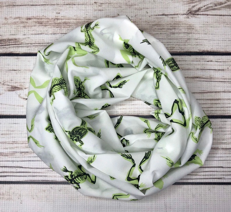 May include: White infinity scarf with a repeating pattern of green skeleton frogs.