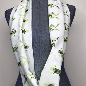 May include: White infinity scarf with a repeating pattern of green frog skeletons.