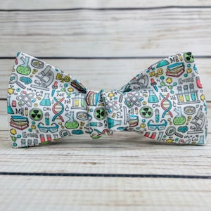 May include: A white bow tie with a science theme. The bow tie is covered in a variety of science-related illustrations, including beakers, test tubes, atoms, molecules, microscopes, and books. The text "H₂O" and "E=mc²" are also visible on the bow tie.