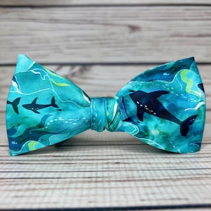 Könnte beinhalten: Eine türkisfarbene Fliege mit einem sich wiederholenden Muster aus schwarzen Haien, die in einem blauen Ozean schwimmen. Die Fliege ist aus Stoff und hat eine klassische Form.