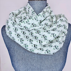 May include: White infinity scarf featuring a repeating pattern of the letters "CTR" in black and green. The scarf is draped over a blue mannequin. This accessory is suitable for various outfits. The scarf's design is clearly visible.