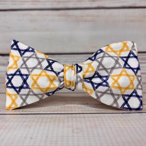 May include: A white bow tie featuring a repeating pattern of blue, yellow, and gray Star of David shapes. The bow tie is made of fabric and is tied in the center. The background is a light wood surface.