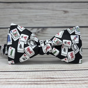 May include: A black bow tie featuring a pattern of white Mahjong tiles with red, green, and blue accents. The bow tie is tied in the center and is set against a light wood grain background.