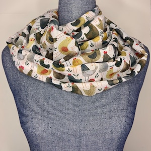 以下が含まれることがあります： 白のインフィニティスカーフ。カラフルな鶏の繰り返し模様が特徴です。鶏は黄色、茶色、グレー、黒など様々な色で、赤い鶏冠と緑の葉のアクセントがあります。スカーフは青いマネキンにかけられています。