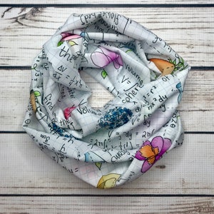 Op de afbeelding: Een witte oneindigheidssjaal met een rasterpatroon en kleurrijke bloemen en wiskundige vergelijkingen erop gedrukt. De vergelijkingen omvatten het volume van een bol, het volume van een kegel en de integraal van een functie.