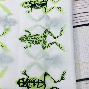 May include: White fabric with a repeating pattern of green frog skeletons. The frogs are in a variety of poses, with their bones visible.