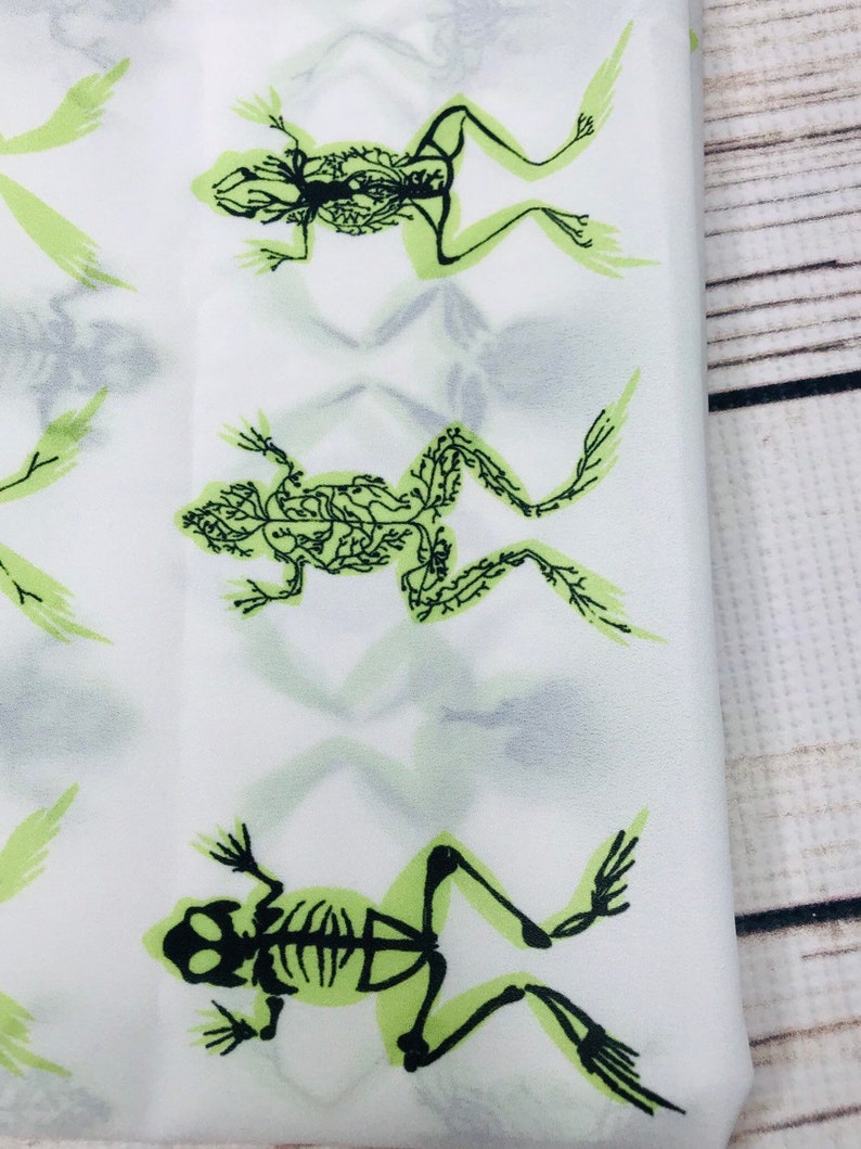 May include: White fabric with a repeating pattern of green frog skeletons. The frogs are in a variety of poses, with their bones visible.