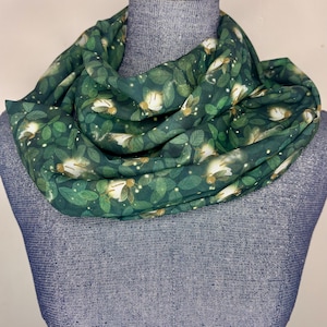 May include: An infinity scarf featuring a dark green background with a repeating pattern of green leaves and glowing fireflies. The fabric has a soft, draped appearance, showcasing the detailed illustrations of the insects and foliage throughout.