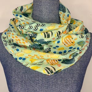 May include: A light green infinity scarf featuring a repeating pattern of various tropical fish in shades of yellow, orange, black, and blue. The fabric is draped in multiple folds, showcasing the colourful aquatic life print across the entire surface.