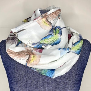 May include: White infinity scarf featuring a colorful hummingbird print. The scarf displays various hummingbirds in shades of blue, green, brown, and red. The scarf is draped over a blue denim form.