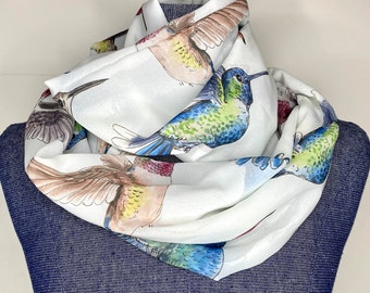May include: White infinity scarf featuring a colorful hummingbird print. The scarf displays various hummingbirds in shades of blue, green, brown, and red. The scarf is draped over a blue denim form.