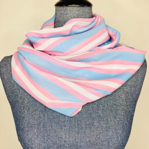 May include: A striped scarf in shades of light blue, pink, and white is draped over a mannequin. The scarf's pattern features alternating horizontal stripes of the three colors. The scarf is made of a soft, lightweight fabric.