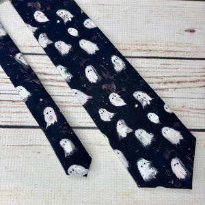 May include: A dark navy blue necktie featuring a pattern of white ghost illustrations. The ghosts have simple, rounded shapes and are scattered across the tie. The tie is made of a smooth fabric and is suitable for Halloween.