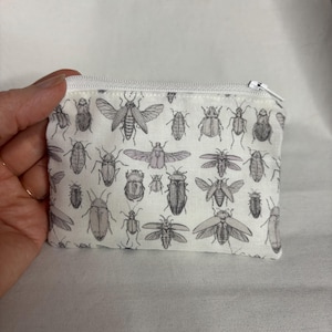 May include: A small, rectangular white pouch with a zipper closure. The pouch is covered in a repeating pattern of detailed, grayscale illustrations of various insects. The zipper is white.