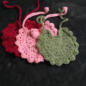 May include: Three handmade crochet bibs in shades of red, pink, and green. Each bib has a scalloped edge and a long, twisted cord for tying. The bibs are arranged on a dark background, showcasing the texture and colors of the yarn.