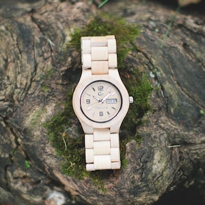May include: A light-colored wooden wristwatch with a matching band. The watch face displays the time and day, with the brand name "Green Forest" visible. The watch is set against a backdrop of moss and wood.