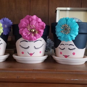 Peut inclure: Trois pots de fleurs décoratifs avec des visages peints et des coiffes florales. Les pots sont noirs avec des visages blancs, des yeux fermés et des accents roses. Chaque pot repose sur une soucoupe assortie. Les coiffes florales sont violettes, roses et turquoise.