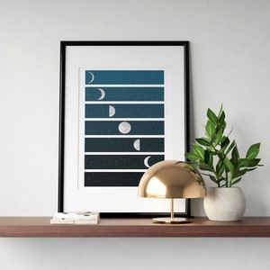 May include: A framed print of a moon phases design with a dark blue background and white stars. The print is on a shelf with a gold lamp and a potted plant.