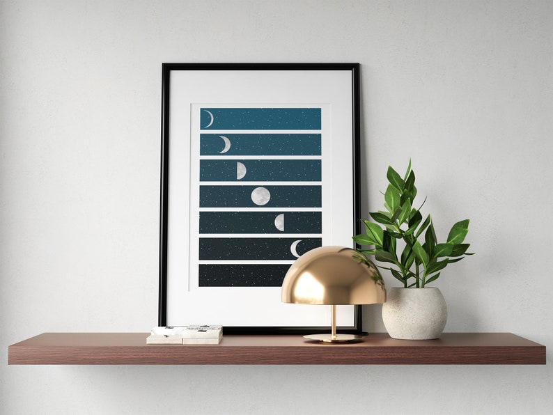 May include: A framed print of a moon phases design with a dark blue background and white stars. The print is on a shelf with a gold lamp and a potted plant.