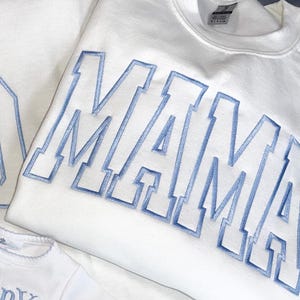 Pode incluir: Close-up de camisolas brancas com a palavra "MAMA" bordada em azul claro. A imagem mostra os detalhes da costura e o tecido macio. Um body branco menor com bordado azul também é visível, sugerindo um conjunto a condizer.