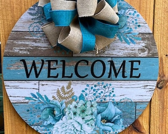 Può includere: Cartello di benvenuto rotondo in legno con sfondo effetto legno invecchiato e decorazioni floreali blu e bianche. La parola "WELCOME" è stampata in nero. Un grande fiocco in juta e nastro turchese è attaccato in alto.