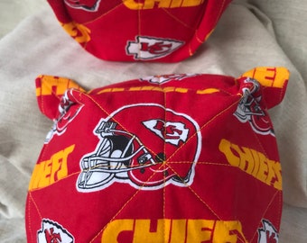 May include: Two red and yellow quilted bowl cozies with the words "CHIEFS" and a helmet design. The bowl cozies have yellow handles and are designed to hold a bowl. One cozy has a white bowl inside.