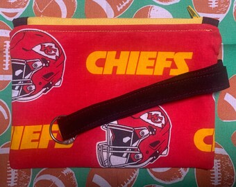 May include: Red zippered pouch with a black strap. The pouch has a repeating pattern of football helmets and the word "CHIEFS" in yellow letters.