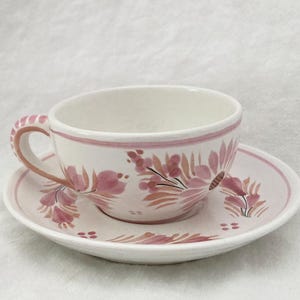 May include: A white ceramic teacup and saucer set decorated with pink floral patterns. The cup has a curved handle and a pink rim. The saucer is round and features the same floral design. The set is suitable for tea or coffee.