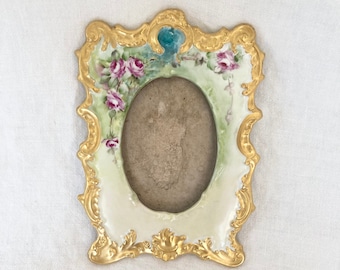 May include: A white porcelain picture frame with a gold ornate design and floral accents. The frame has an oval opening for a photo.