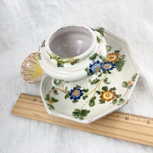 May include: A white ceramic dish with a floral design in shades of blue, yellow, and green. The dish has a raised center with a small, round, white ceramic pot with a lid. The pot has a floral design similar to the dish.