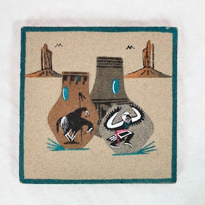 Puede incluir: Una pintura de arena que representa a un bailarín nativo americano con un traje marrón y blanco con una faja roja, rodeado de dos edificios marrones y un caballo marrón con un jinete. El fondo es una escena desértica de color beige con dos formaciones rocosas marrones, altas y delgadas.