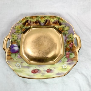 May include: A square porcelain serving dish with two handles. The dish is decorated with a gold rim and a painted scene of a woodland with berries and plums.
