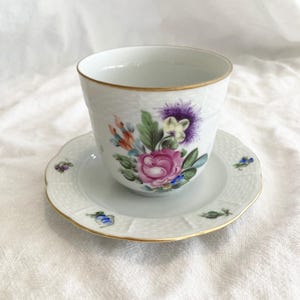 Może przedstawiać: Zestaw filiżanki i spodeczka do herbaty z białej porcelany z kwiatowym wzorem. Filiżanka i spodeczek mają złotą obwódkę i delikatny kwiatowy wzór w odcieniach różowego, fioletowego, niebieskiego i zielonego.