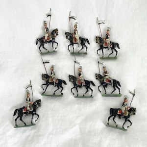 May include: A group of antique toy soldiers riding black horses. Each soldier is dressed in a uniform and holds a small flag. The figures are painted with various colours and are set on green bases.