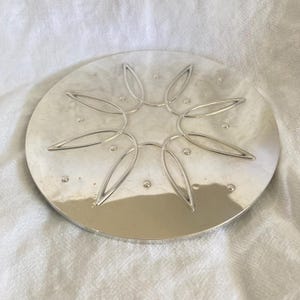 May include: A round, silver-colored trivet with a floral design in the center. The trivet has a series of small, evenly spaced dots around the edge. The floral design is made of curved, petal-like shapes.