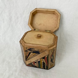 Könnte beinhalten: Geöffnete, achteckige Holzkiste mit Scharnierdeckel. Die Kiste ist mit einem geometrischen Design in verschiedenen Farben verziert. Der Deckel ist geöffnet und gibt den Blick ins Innere frei. Das Äußere weist eine Mischung aus Formen und Farben auf, darunter Blau, Rot und Gelb.
