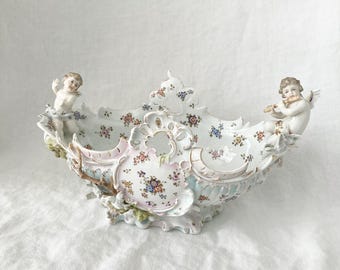 Puede incluir: Un cuenco central decorativo de porcelana con un diseño floral y dos figuras de querubines. El cuenco presenta una paleta de colores pastel con acentos rosas, azules y dorados. Los querubines son blancos y detallados.