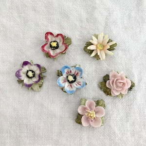 Puede incluir: Seis adornos de flores de porcelana con hojas verdes. Las flores son de varios colores, incluyendo rosa, azul, morado y rojo.