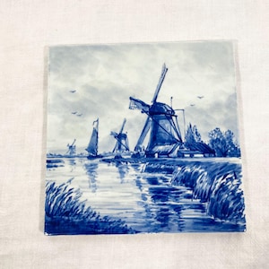 Può includere: Una piastrella di Delft blu e bianca che raffigura un paesaggio olandese con mulini a vento e un canale. La piastrella è dipinta a mano e presenta un design tradizionale.